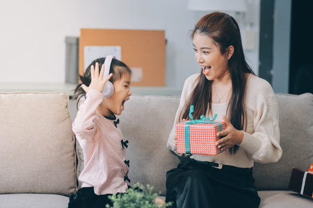 A cheerful Asian mother helps her young daughter do homework and color a book at home, while the father works on a laptop in the background. A cozy, bright, and modern family lifestyle.の写真素材