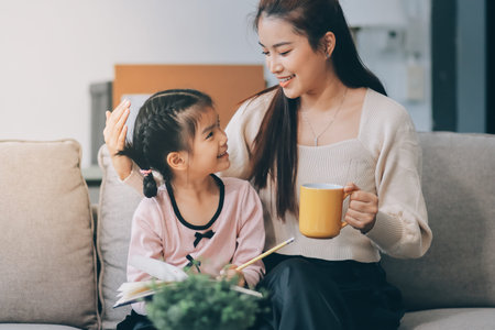 A cheerful Asian mother helps her young daughter do homework and color a book at home, while the father works on a laptop in the background. A cozy, bright, and modern family lifestyle.の写真素材