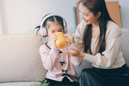 A cheerful Asian mother helps her young daughter do homework and color a book at home, while the father works in the background. A cozy, bright, and modern family lifestyle.の写真素材