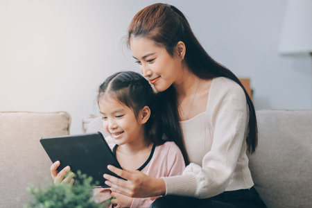 A cheerful Asian mother helps her young daughter do homework and color a book at home, while the father works on a laptop in the background. A cozy, bright, and modern family lifestyle.の写真素材