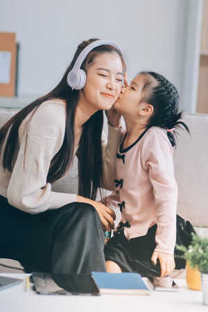 A cheerful mother helps her young daughter do homework and color a book at home, while the father works on a laptop in the background. A cozy, bright, and modern family lifestyle.の写真素材