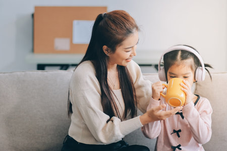 A cheerful Asian mother helps her young daughter do homework and color a book at home, while the father works on a laptop in the background. A cozy, bright, and modern family lifestyle.の写真素材