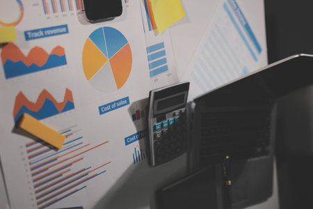 business documents on office table with tablet, smart phone and laptop and two colleagues discussing dataの写真素材