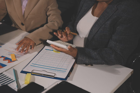 business documents on office table with tablet, smart phone and laptop and two colleagues discussing dataの写真素材