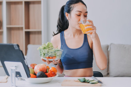 Fit young Asian women in activewear sitting on yoga mat and eating fresh vegetable salad after...
