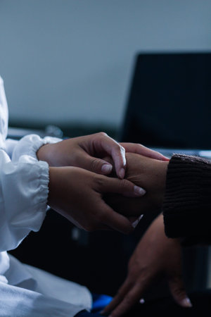 Senior patient lying in hospital bed receiving comforting touch from young female nurse, hands gently clasped together, medical care setting visibleの写真素材