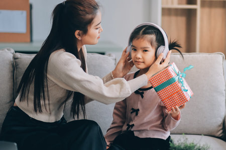 A cheerful Asian mother helps her young daughter do homework and color a book at home, while the father works on a laptop in the background. A cozy, bright, and modern family lifestyle.の写真素材