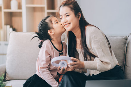 A cheerful Asian mother helps her young daughter do homework at home, while the father works on a laptop in the background. A cozy, bright, and modern family lifestyle.の写真素材