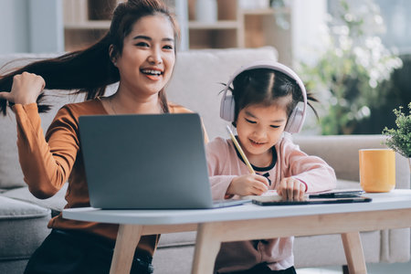 A cheerful Asian mother helps her young daughter do homework and color a book at home, while the father works on a laptop in the background. A cozy, bright, and modern family lifestyle.の写真素材