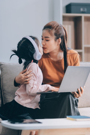 A cheerful Asian mother helps her young daughter do homework and color a book at home, while the...