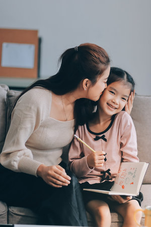 A cheerful Asian mother helps her young daughter do homework and color a book at home, while the father works on a laptop in the background. A cozy, bright, and modern family lifestyle.の写真素材