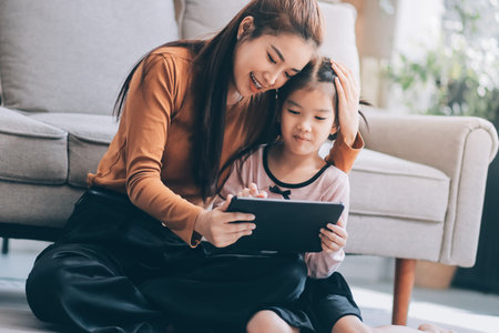 A cheerful Asian mother helps her young daughter do homework and color a book at home, while the father works on a laptop in the background. A cozy, bright, and modern family lifestyle.の写真素材