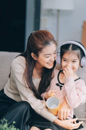 A cheerful Asian mother helps her young daughter do homework and color a book at home, while the father works on a laptop in the background. A cozy, bright, and modern family lifestyle.の写真素材