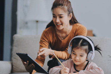A cheerful Asian mother helps her young daughter do homework and color a book at home, while the father works on a laptop in the background. A cozy, bright, and modern family lifestyle.の写真素材
