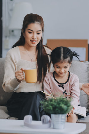 A cheerful Asian mother helps her young daughter do homework and color a book at home, while the...
