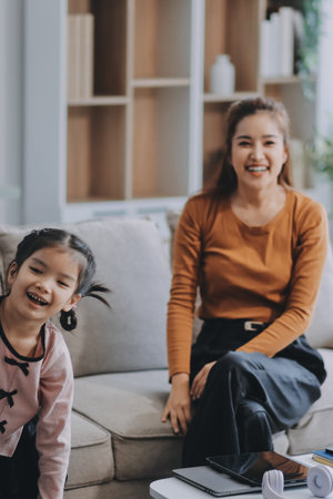 A cheerful Asian mother helps her young daughter do homework and color a book at home, while the...