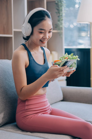 Fit young Asian women in activewear sitting on yoga mat and eating fresh vegetable salad after...