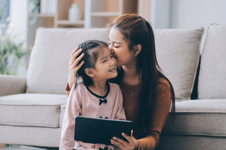 A cheerful Asian mother helps her young daughter do homework and color a book at home, while the...
