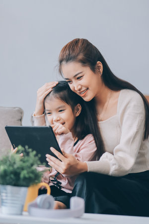 A cheerful Asian mother helps her young daughter do homework and color a book at home, while the...