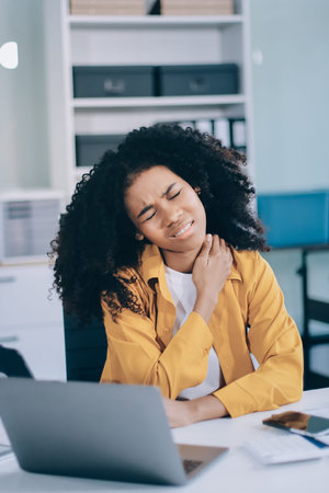 Businesswoman experiencing discomfort and pain in her neck while sitting at her desk, working on a...