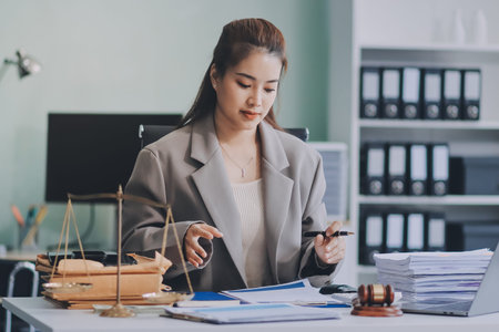 Confident Lawyer Working at Desk: A young Asian female lawyer confidently works on her laptop, surrounded by legal symbols. She exudes professionalism and competence in her modern office setting.の写真素材