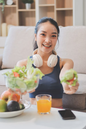 Fit young Asian women in activewear sitting on yoga mat and eating fresh vegetable salad after...