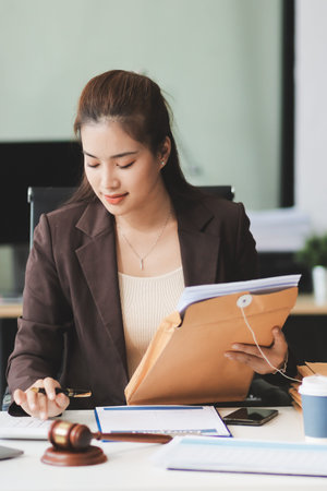 Confident Lawyer Working at Desk: A young Asian female lawyer confidently works on her laptop, surrounded by legal symbols. She exudes professionalism and competence in her modern office setting.の写真素材