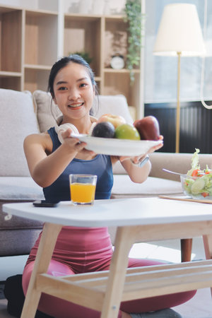 Fit young Asian women in activewear sitting on yoga mat and eating fresh vegetable salad after...