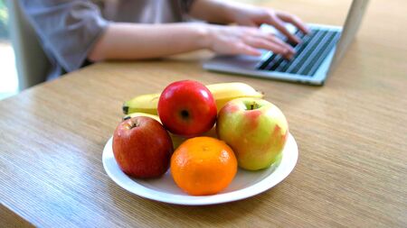 A plate of fruits on a table with the hands of a woman on a laptop in backgroundの写真素材