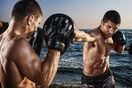 Fighter is training his boxing skills with a coach. Outdoor training on a beach. Boxing, mma, wrestling, karate. Young, strong, muscular athleteの写真素材