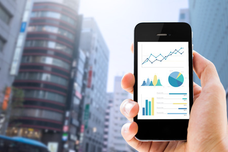 Close-up of smartphone showing graphs,charts and diagrams on screen.  Male's hand using it  against blurred blue modern buildings and sky background. cityscape view. planning and analysis concept.の写真素材
