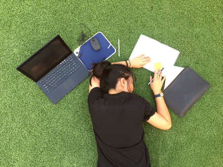 Working woman stressed out from work and holding the head with lay down on green grass with laptop and notebook.の写真素材