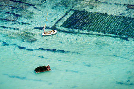 view of the coast with beach and coral at bukit in bali -farmer in boat harvesting sea weed in underwater plantation-indonesiaの写真素材