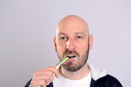 bald headed man brushing teeth in front of gray background