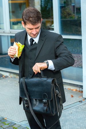 businessman in suit in front of city- background is eating and looking for timeの写真素材