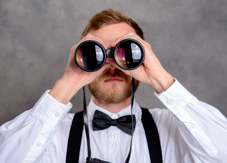 young bearded man in white shirt and bow tie looking through a binocularsの写真素材