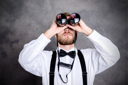 young bearded man in white shirt and bow tie looking through a binocularsの写真素材