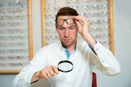 in optician shop- young man with magnifier selecting new reading glasses