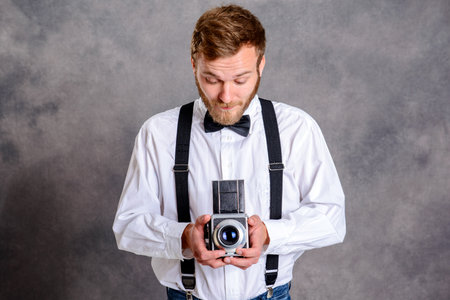 young bearded man in white shirt and bow tie with old camera standing in front of gray backgroundの写真素材