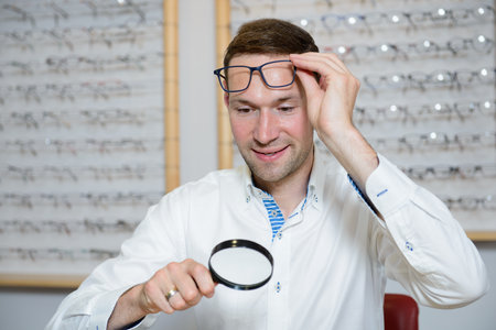 in optician shop- young man with magnifier selecting new reading glassesの写真素材
