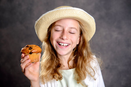 smiling girl with blond hair in white dress and straw hat eating muffinの写真素材