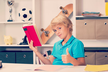 blond boy with thumb up reading a bookの写真素材