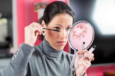 young dark haired woman looking in mirror and applying make upの写真素材