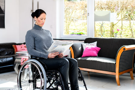 young disabled woman in wheelchair at home reading newspaper and drinking coffeeの写真素材