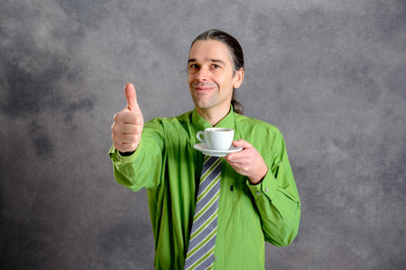 young man in green shirt and necktie drinking coffee and showing thumb upの写真素材