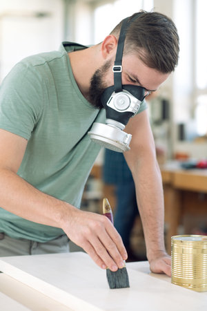 young worker with workpiece in a carpenter's workshop painting wood with brush and respiratorの写真素材