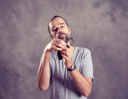 young bearded man ceaning his glasses in front of gray backgroundの写真素材