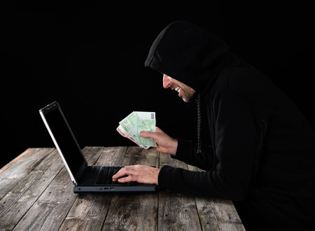 hacker in black hoody with laptop and euro notes in front of black background