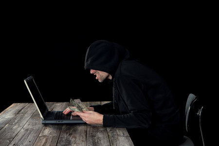hacker in black hoody with laptop and dollar notes in front of black backgroundの写真素材