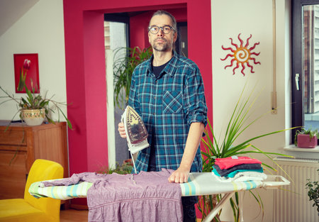 man with iron on ironing board with clothers in living roomの写真素材
